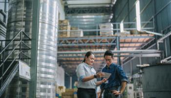 An Image Of Two People Looking At A Tablet In A Factory Setting, Surrounded By Storage Such As Pallet Racks.